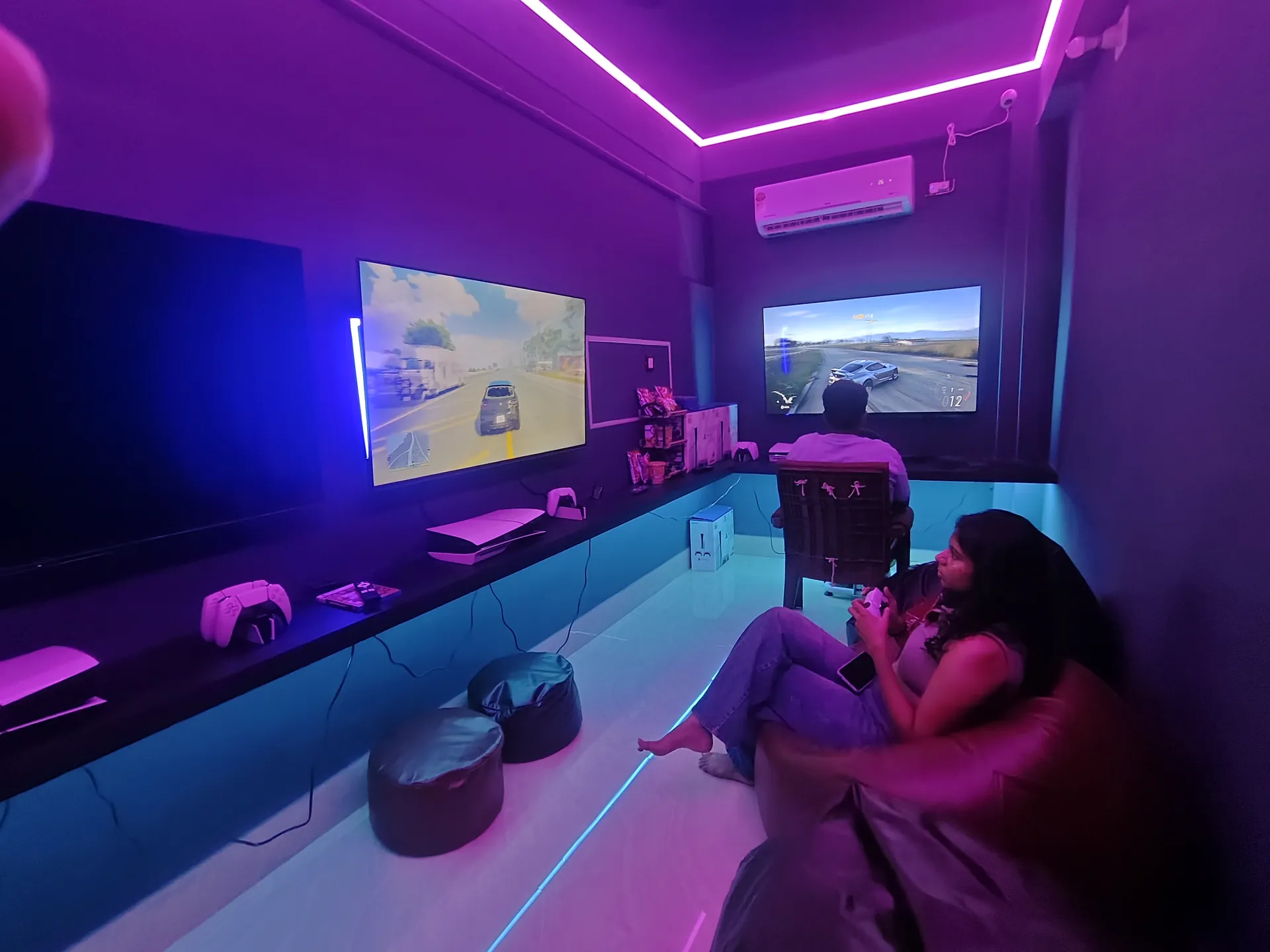 Two players gaming on PS5 with a wall-mounted display and ambient neon lighting